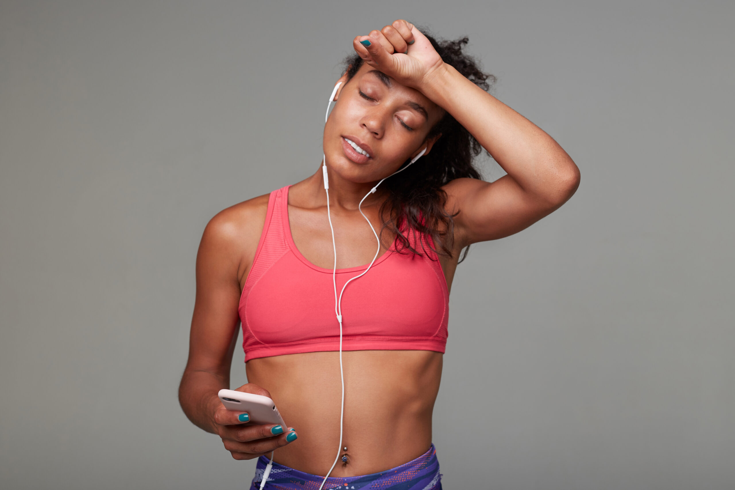 Good looking young fitness dark skinned brunette model wearing earphones while posing over grey background, wiping sweat from forehead with hand and keeping eyes closed